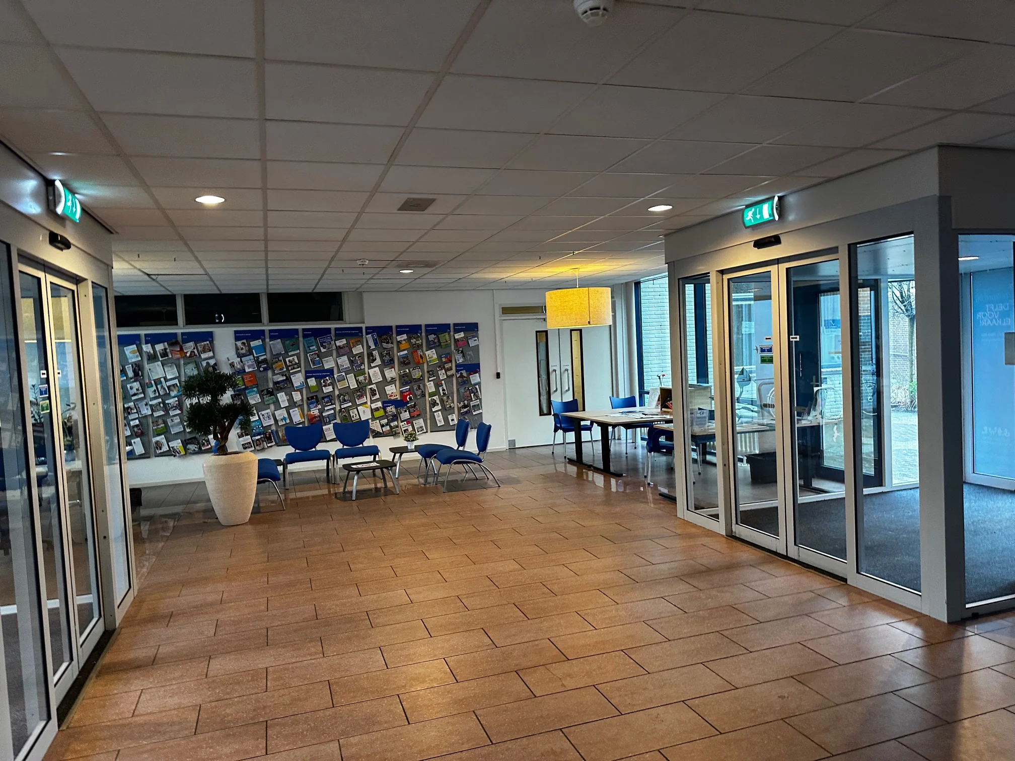 Hal met glazen deuren, blauwe stoelen en een wand met brochures in een gebouw aan de Van Bleyswijckstraat.